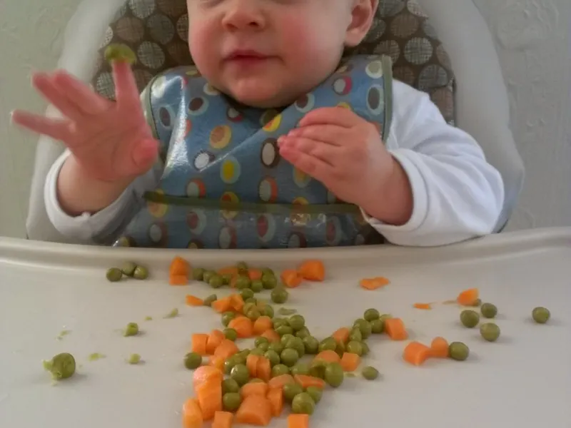 Baby in a highchair eating peas and carrots from the tray